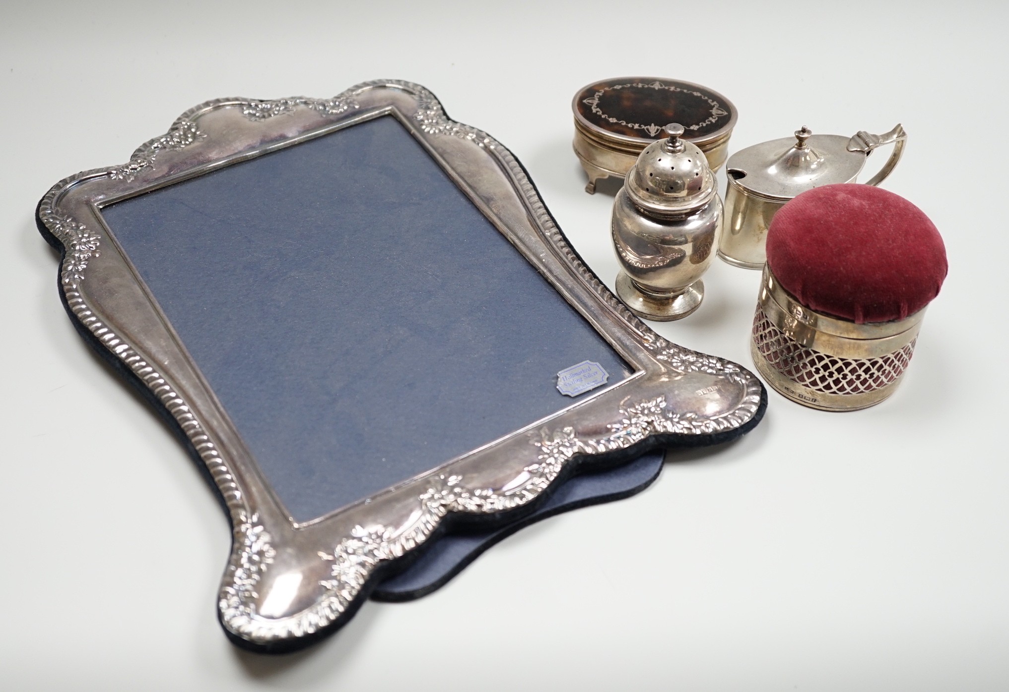 A modern silver mounted photograph frame, 30.2cm, an Edwardian silver mounted pin cushion, mustard pot, salt and a George V silver and tortoiseshell pique mounted trinket box.